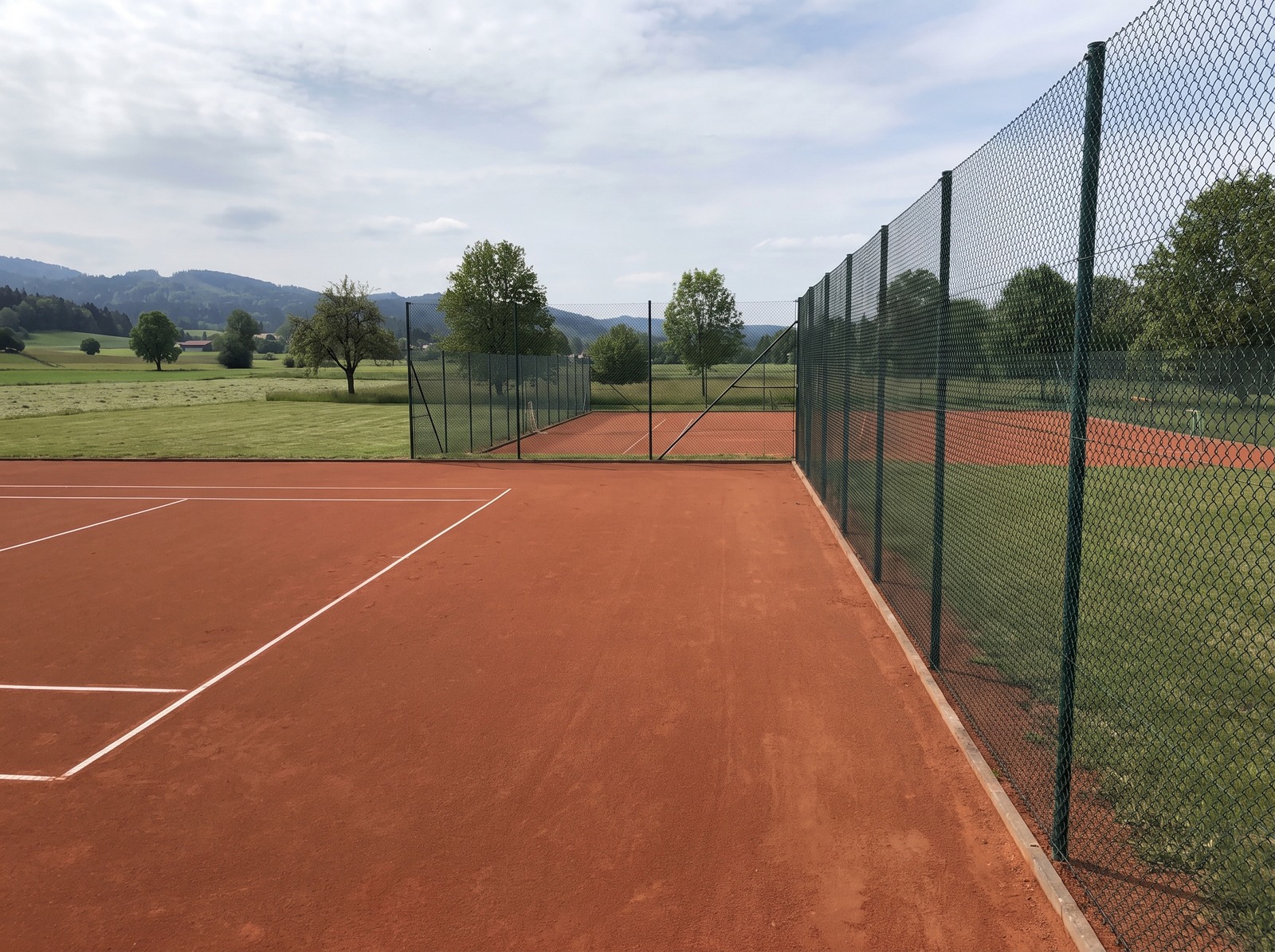 Sandplatz einer neuen Tennisanlage mit Ballfangzaun, dahinter ein zweiter Platz